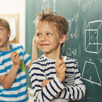 Cute little boys in math class, smiling back to the teacher. There are mathematic symbols written at the brackboard.