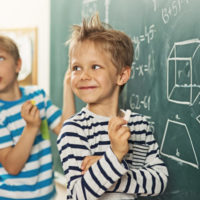 Cute little boys in math class, smiling back to the teacher. There are mathematic symbols written at the brackboard.