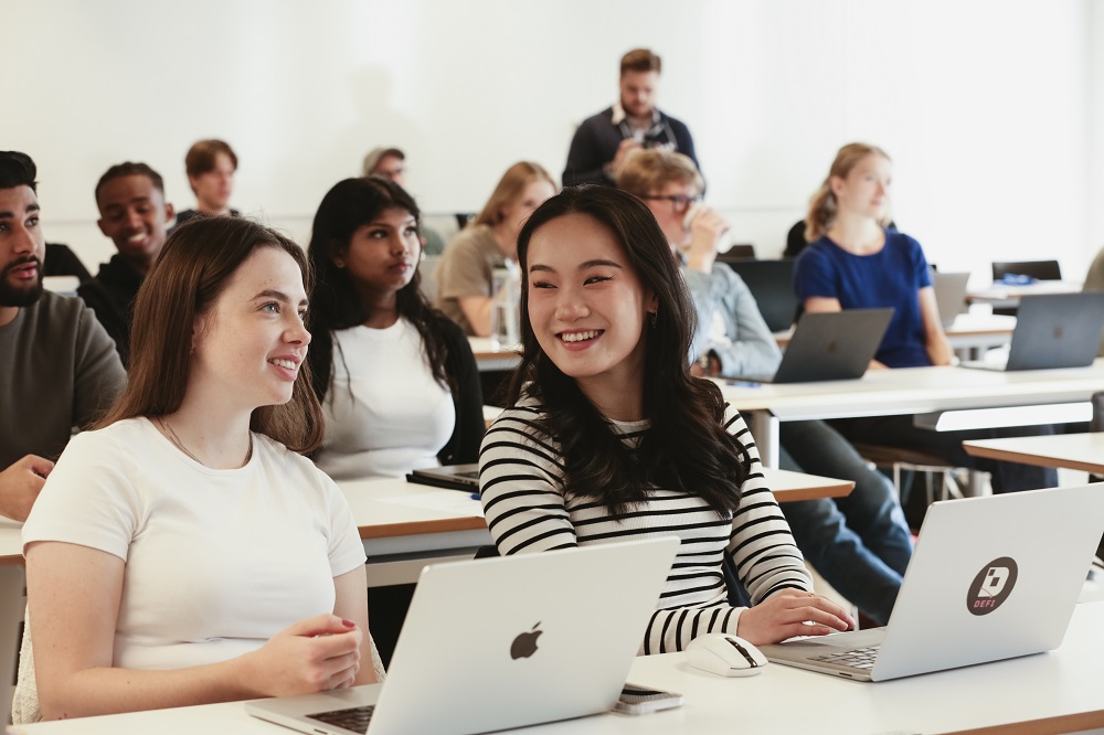 Two higher ed students smiling with other students seated behind them in a classroom
