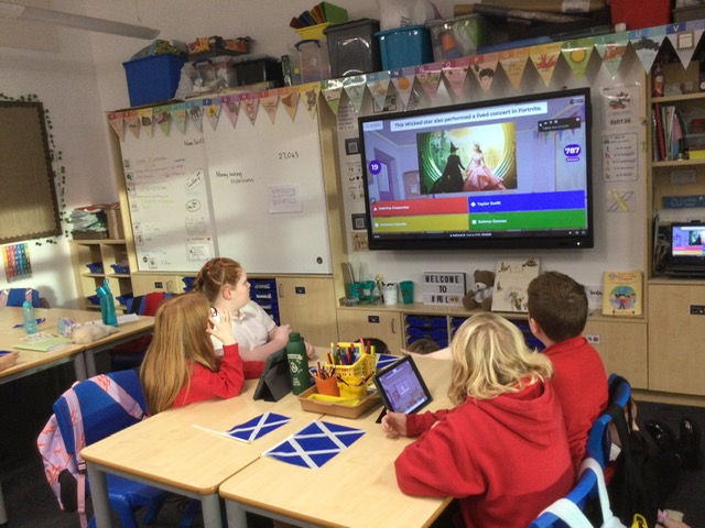 4 students sitting at a cohort of desks in a classroom, watching a large screen with a Zoom Kahoot! presented