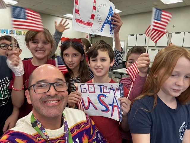 teacher Steve with students in background, waving USA flags and smiling for a selfie photo