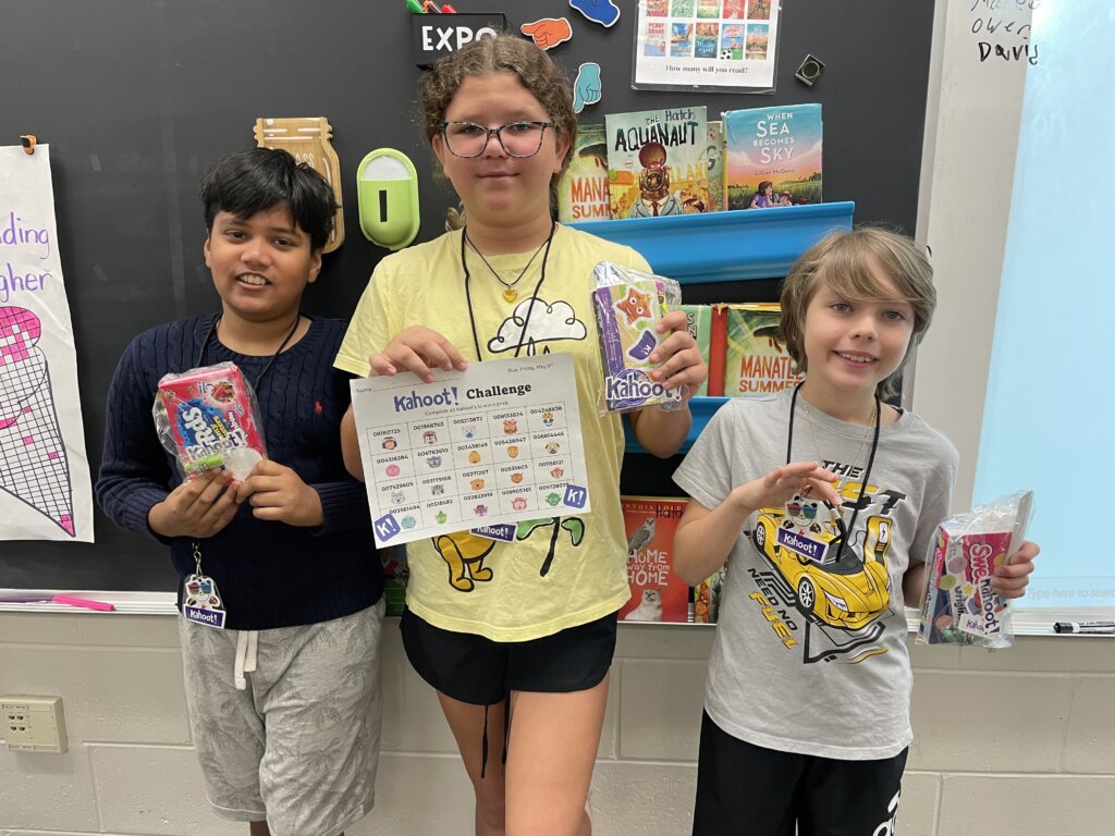 three students smiling at camera holding prizes and kahoot game board