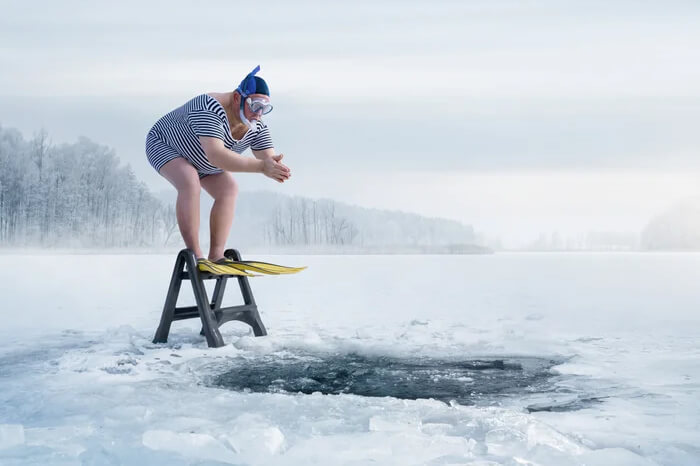 Humorous image of a person in a striped swimsuit, snorkel mask, and flippers preparing to jump into a hole cut in the ice of a frozen lake.