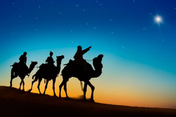 Silhouette of the Three Kings (Magi) riding camels across a desert dune at twilight, following the bright Star of Bethlehem in the night sky.