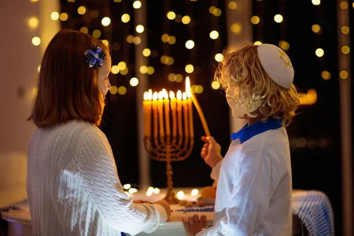 Two young children, one wearing a kippah, lighting the candles on a menorah for the Jewish holiday of Hanukkah, with bokeh lights in the background.