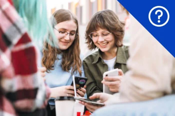 Two smiling young women wearing glasses and holding smartphones, sitting at a table with friends, with a large blue question mark icon overlaid in the corner.