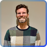 A headshot of a smiling man with brown hair, a beard, and glasses. He is wearing a colorful, checkered knit sweater in shades of green, tan, navy, and white. The photo is set against a plain beige background and framed by a thin, blue-to-gray gradient border.