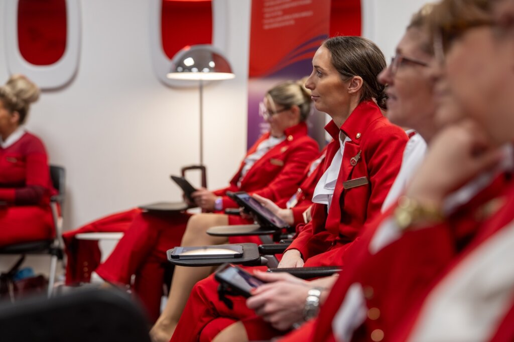 A group of Virgin Atlantic flight attendants are paying attention to a trainer in a classroom setting.