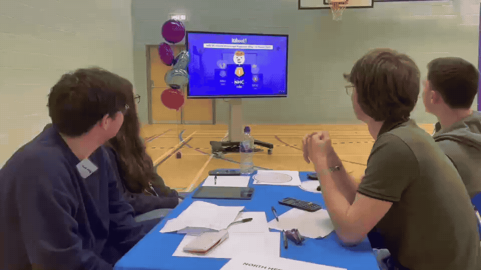Group of 4 students around a working table with kahoot podium screen and animation playing in background