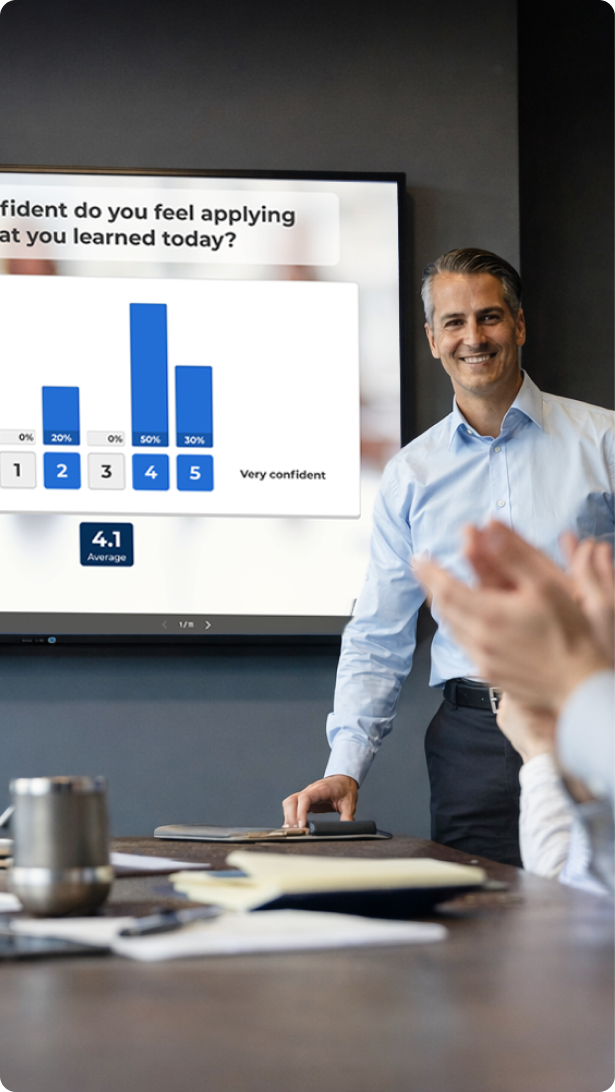 A smiling presenter in a light blue shirt stands in a conference room as an audience member in the foreground claps.