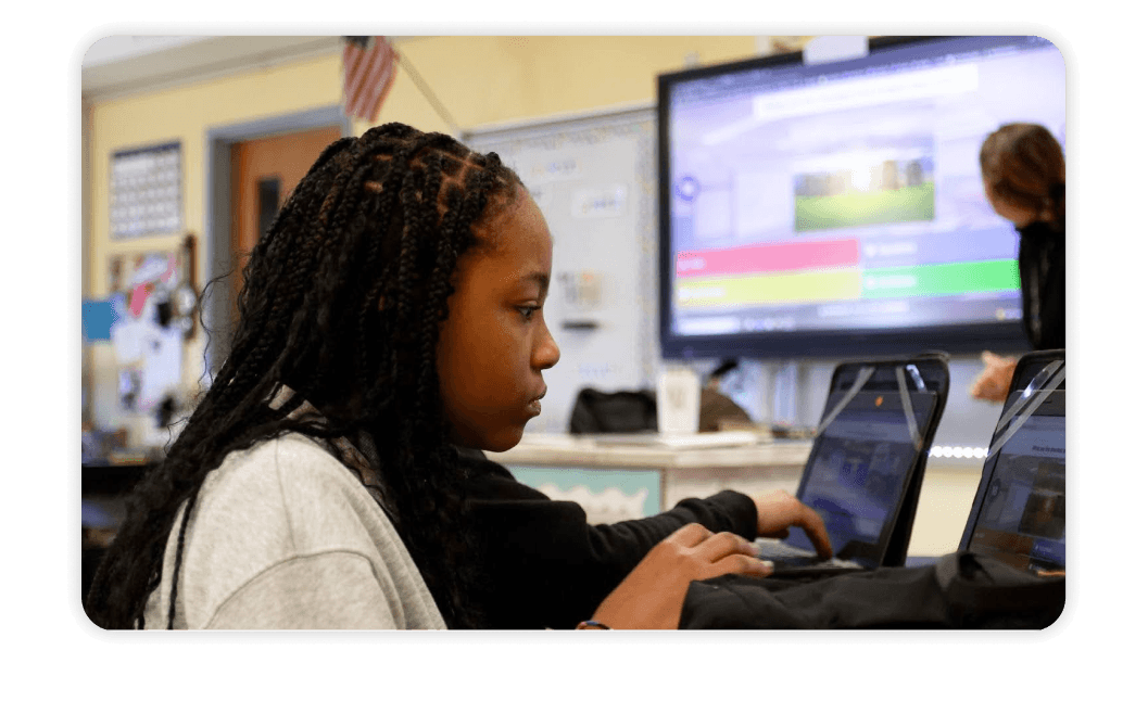 A middle school student with braids focuses on a laptop during a Kahoot! quiz, with a classroom display screen showing the game in the background.