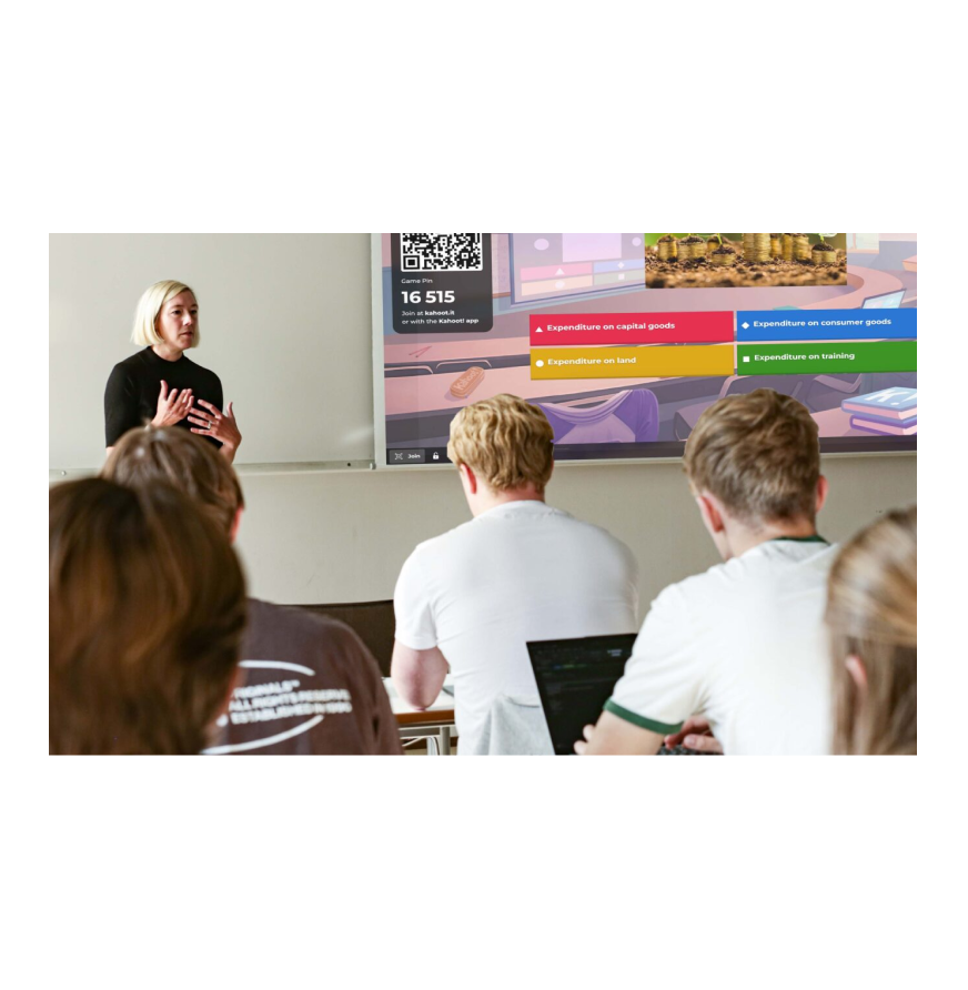 A university lecturer presents a Kahoot! quiz to students in a lecture hall, with the game displayed on a large screen behind her.
