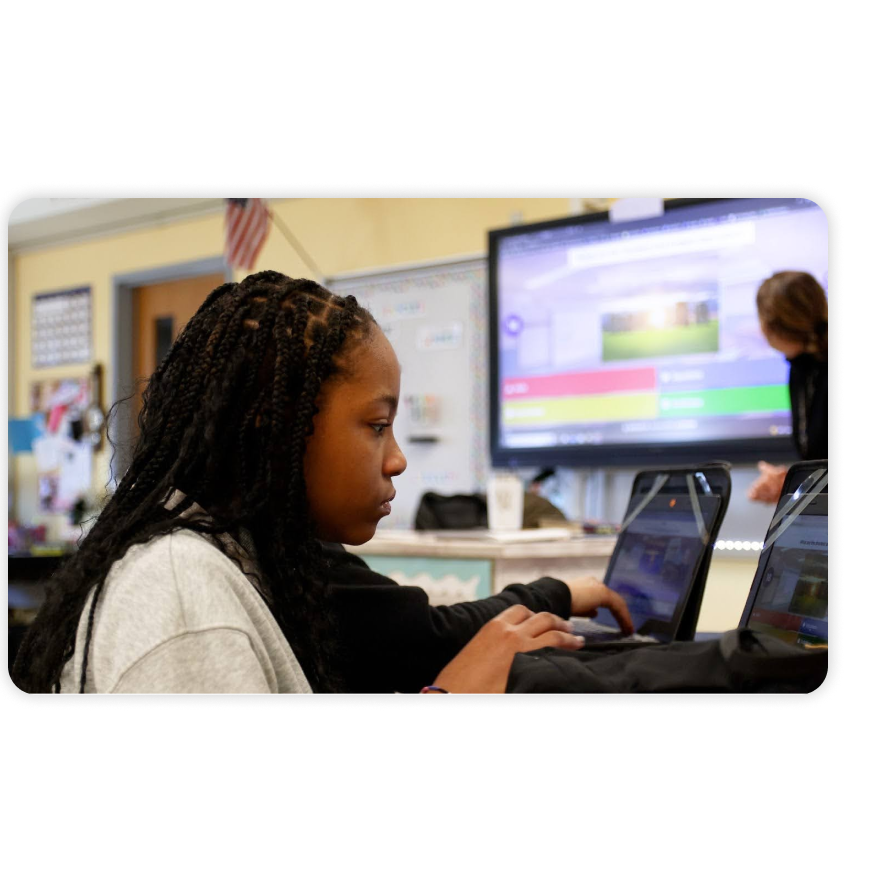 A student focuses on a laptop during a Kahoot! quiz, with a classroom display screen showing the game in the background.