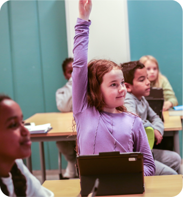 Young girl raising her hand enthusiastically in a classroom with a tablet on her desk