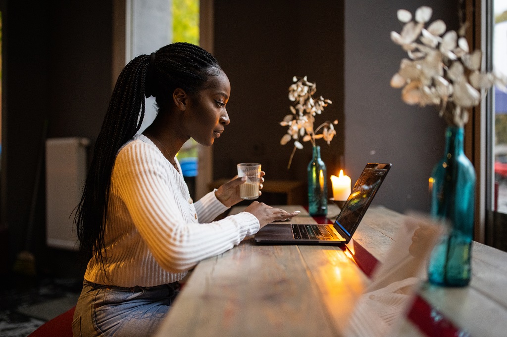 Woman at coffee shop working on laptop. Side view of african female holding a cup of coffee and using laptop at cafe.