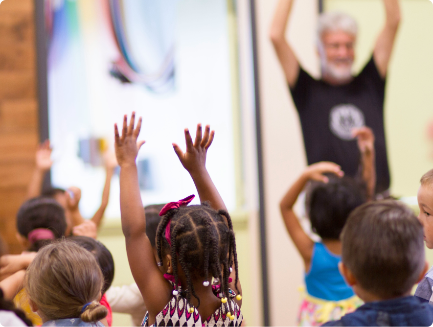Young children enthusiastically raising their hands during a classroom activity with a teacher at the front