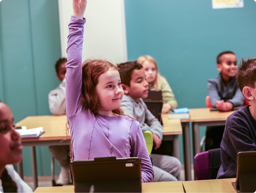 Young girl raising her hand in a classroom while other students with laptops look on