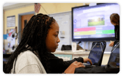 A middle school student with braids focuses on a laptop during a Kahoot! quiz, with a classroom display screen showing the game in the background.