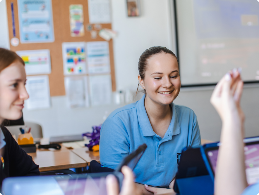 Two students in school uniforms smiling in class, one raising their hand with a tablet visible