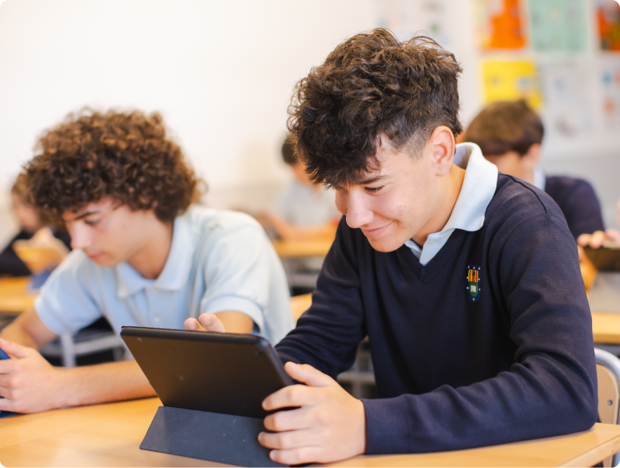 Male student in school uniform smiling while using a tablet at his classroom desk