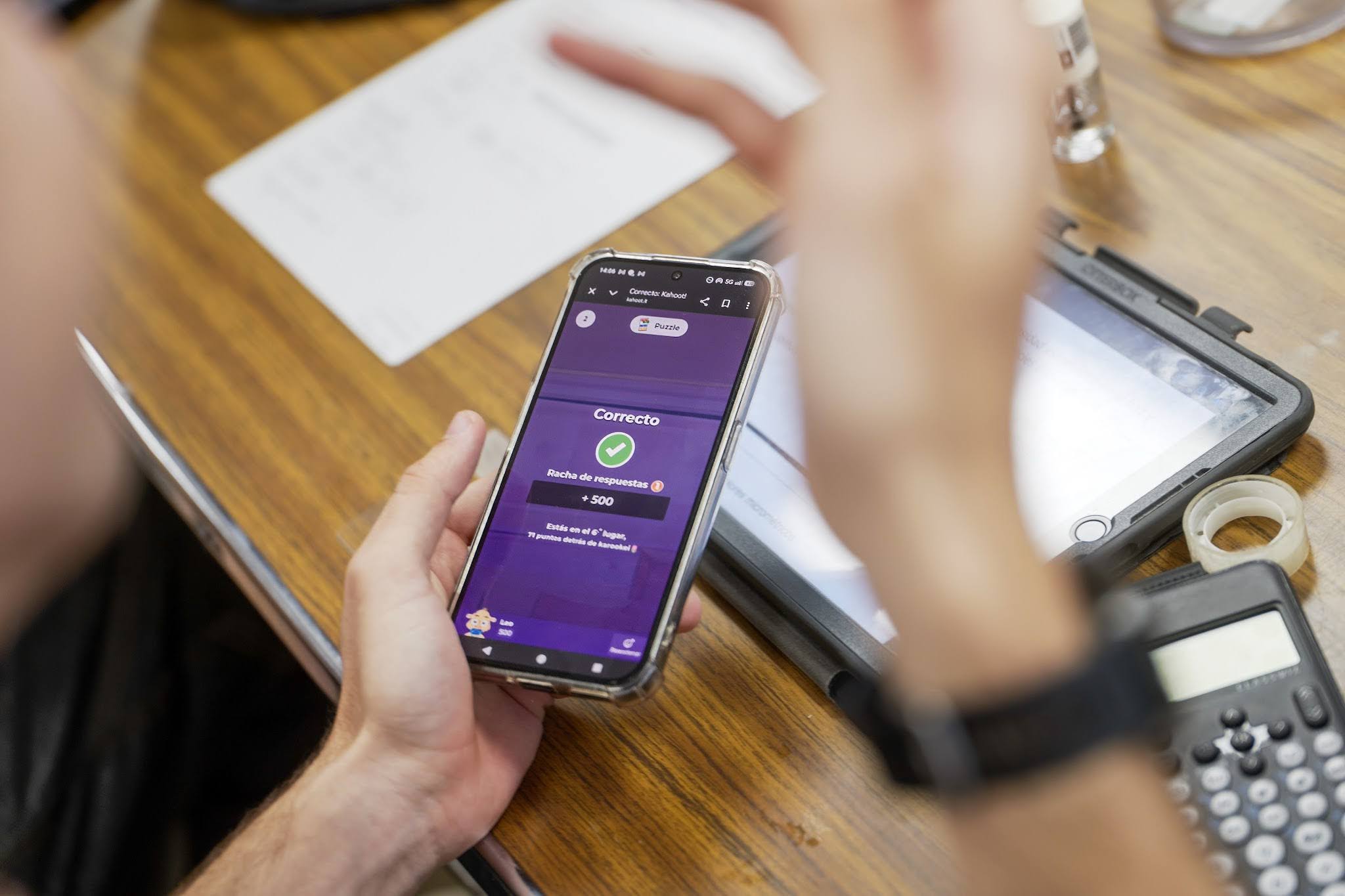 A photo of a mobile phone displaying a kahoot session, held in a person's hand over a desk.
