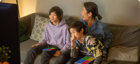 A mother and two children sit on a couch smiling while playing Kahoot together on their devices.