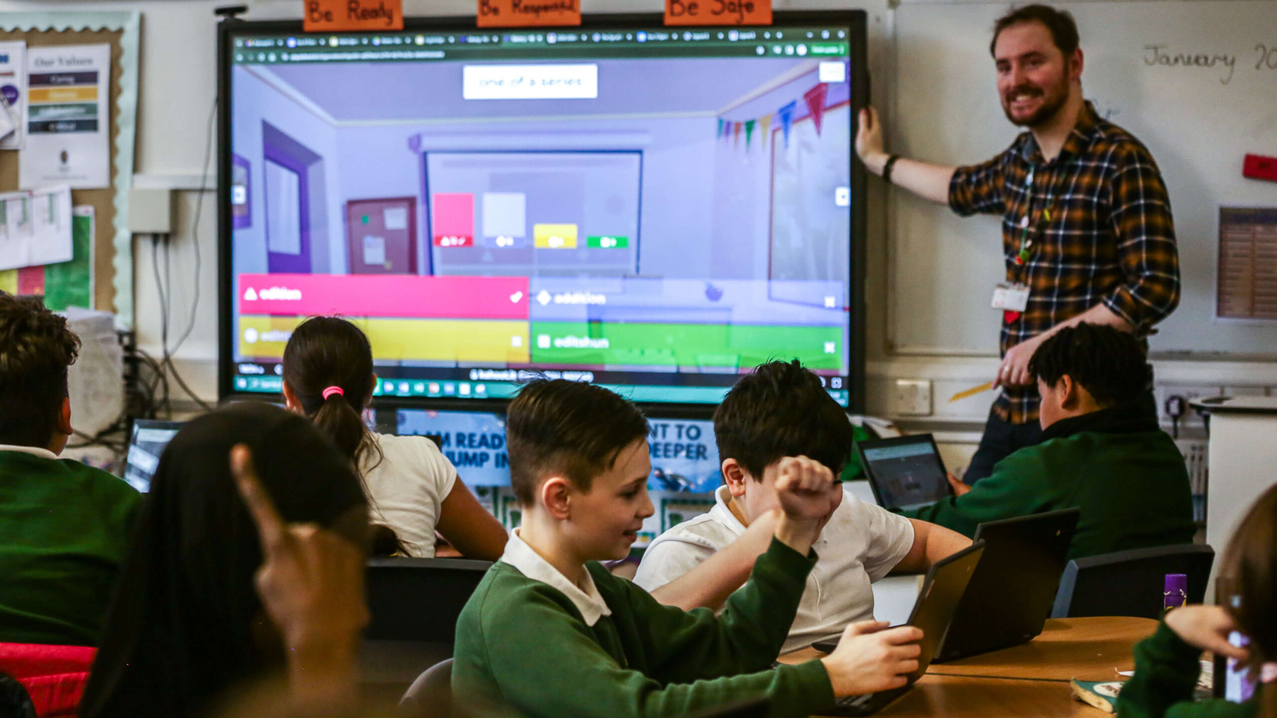 A teacher smiles at the front of a classroom next to a large interactive display showing a Kahoot! quiz, while students use laptops at their desks and one student enthusiastically raises his fist.