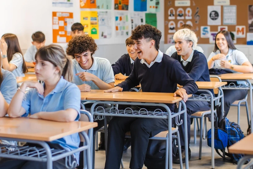A group of secondary school students in uniform laugh and chat together at their desks during class.