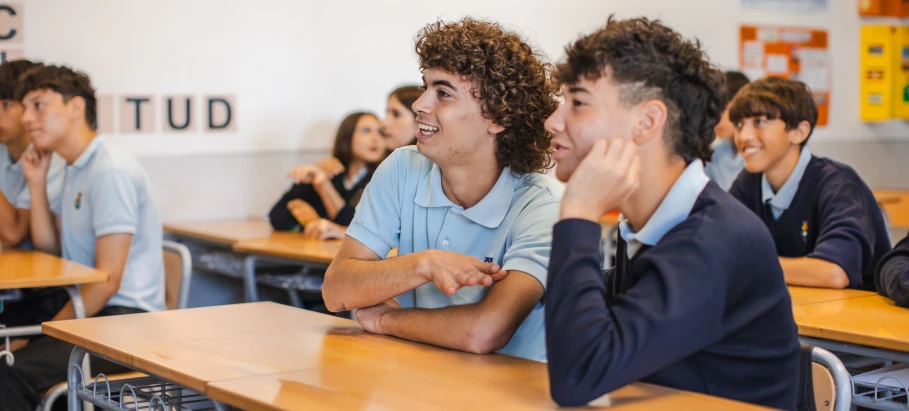 Two secondary school students in uniform sit at desks smiling and chatting during a classroom lesson.