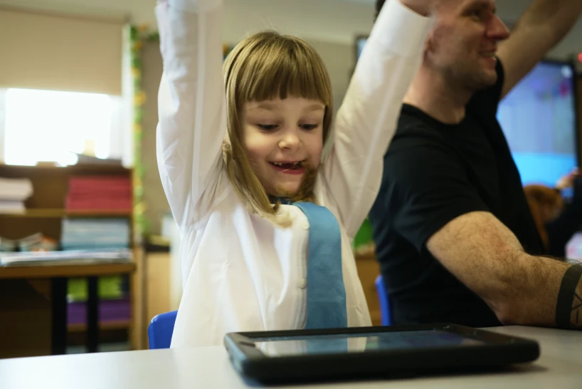 A young student throws her arms up in celebration while playing Kahoot on a tablet, with an adult beside her doing the same.