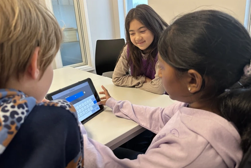 Three young students gather around a tablet, smiling as one of them types into a Kahoot game.