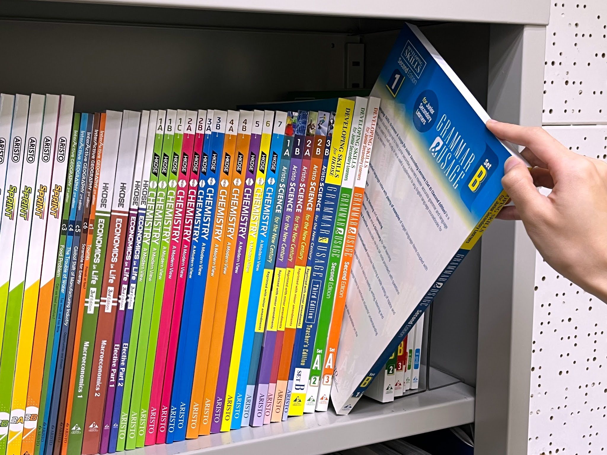 A photo of a hand pulling a grammar textbook from a full book shelf of textbooks