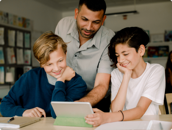 A teacher leans over two smiling students as they look at a tablet together in a classroom.