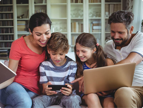 A smiling family of four sits together on a sofa, playing on a smartphone, tablet and laptop.